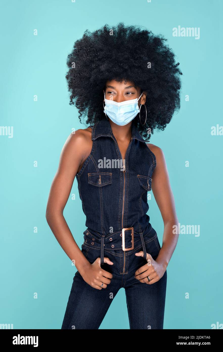 Happy African American femmina in abiti alla moda denim e maschera viso guardando lontano mentre in piedi su sfondo turchese durante pandemic Foto Stock