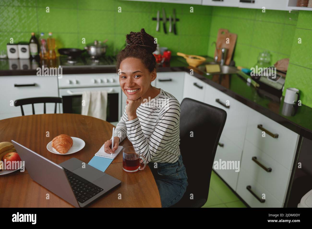 Una bella donna di razza mista che lavora su un notebook e scrive in un notebook a casa al chiuso Foto Stock