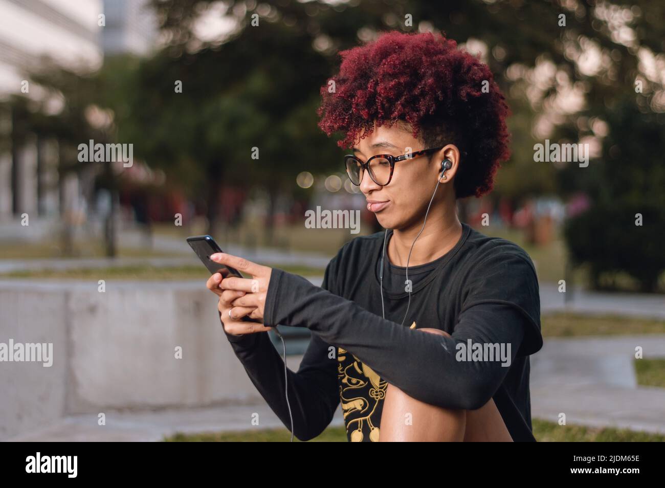 vista laterale, bella giovane donna bruna latina con afro, sorridente felice seduta all'aperto controllare il suo telefono, controllare le notifiche sul social network Foto Stock