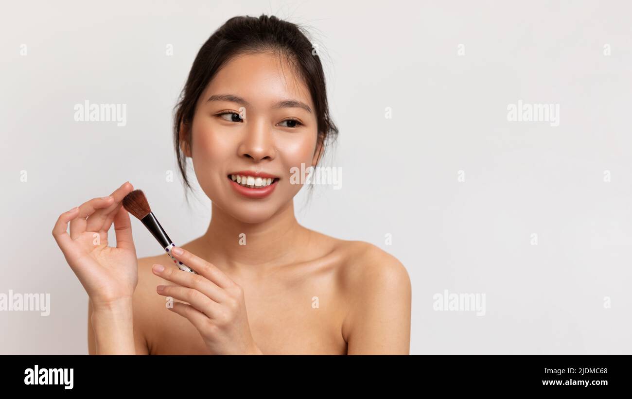Concetto di trucco giornaliero. Ritratto di giovane donna asiatica con la bellezza naturale che tiene pennello Blusher, posando su sfondo bianco Foto Stock