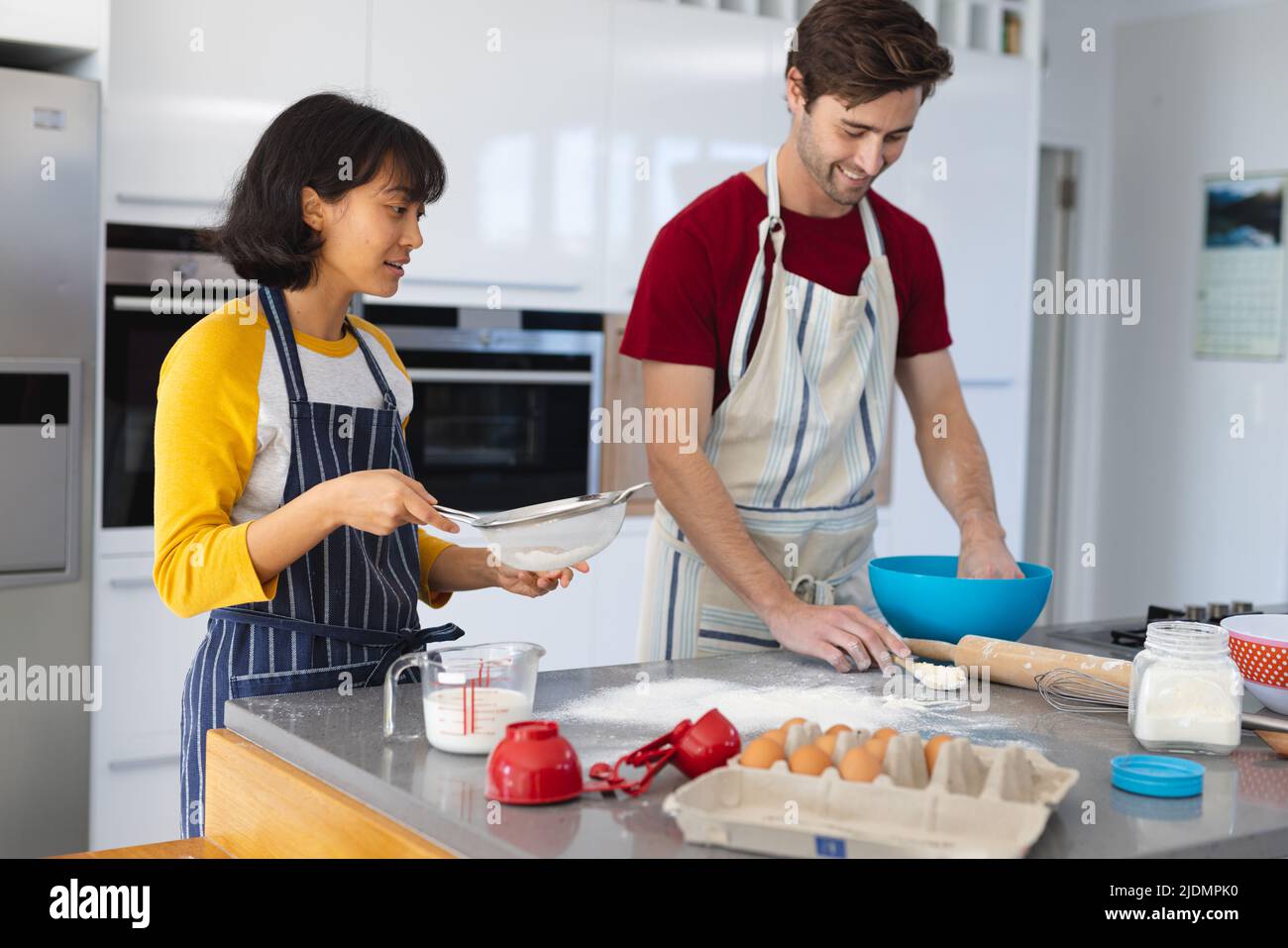 Giovane caucasico che mescola pastella in ciotola mentre donna asiatica che tiene il setaccio all'isola della cucina Foto Stock