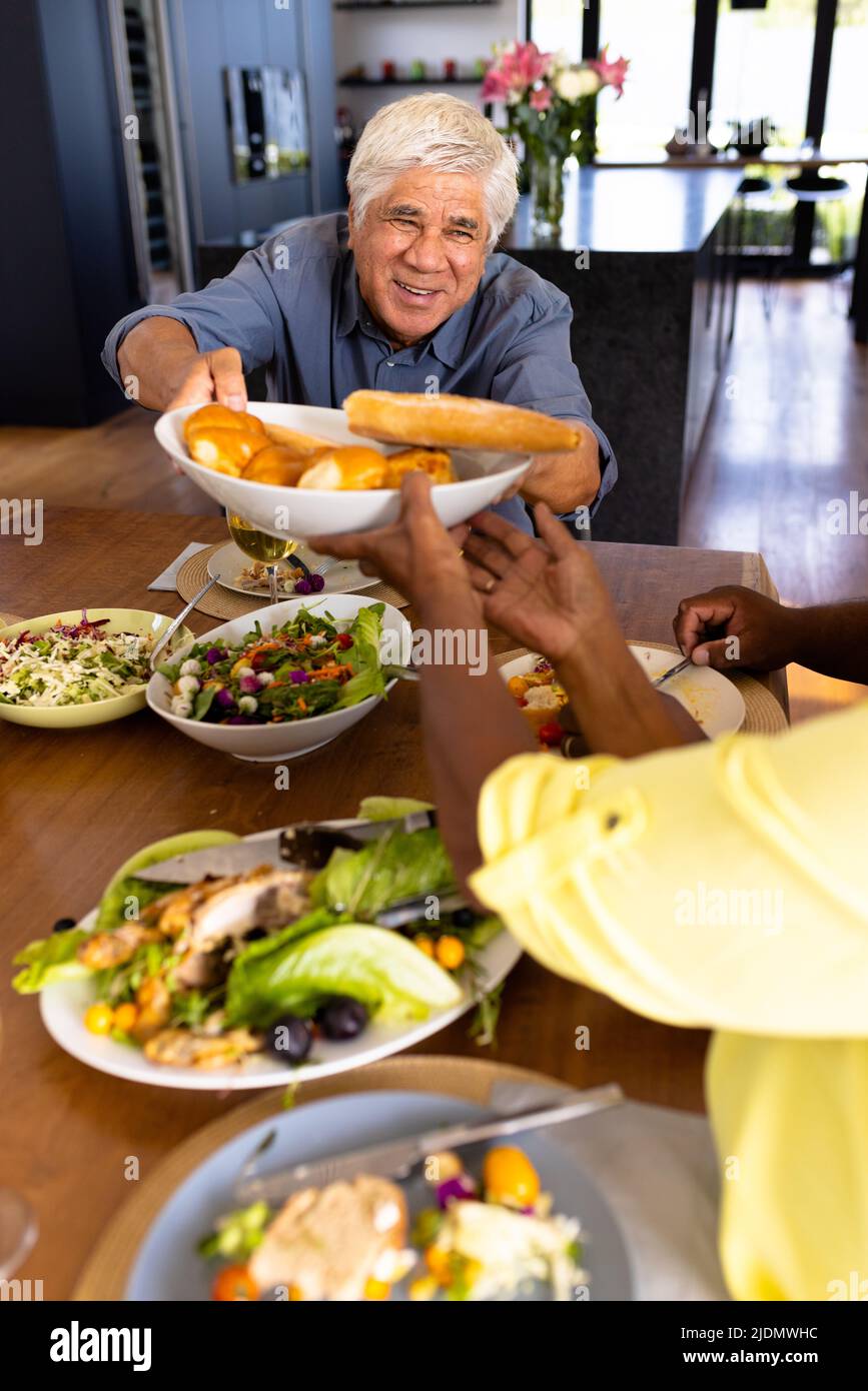 Sorridente uomo anziano multirazziale che dà il pane ad un amico femminile mentre pranzano al tavolo da pranzo Foto Stock