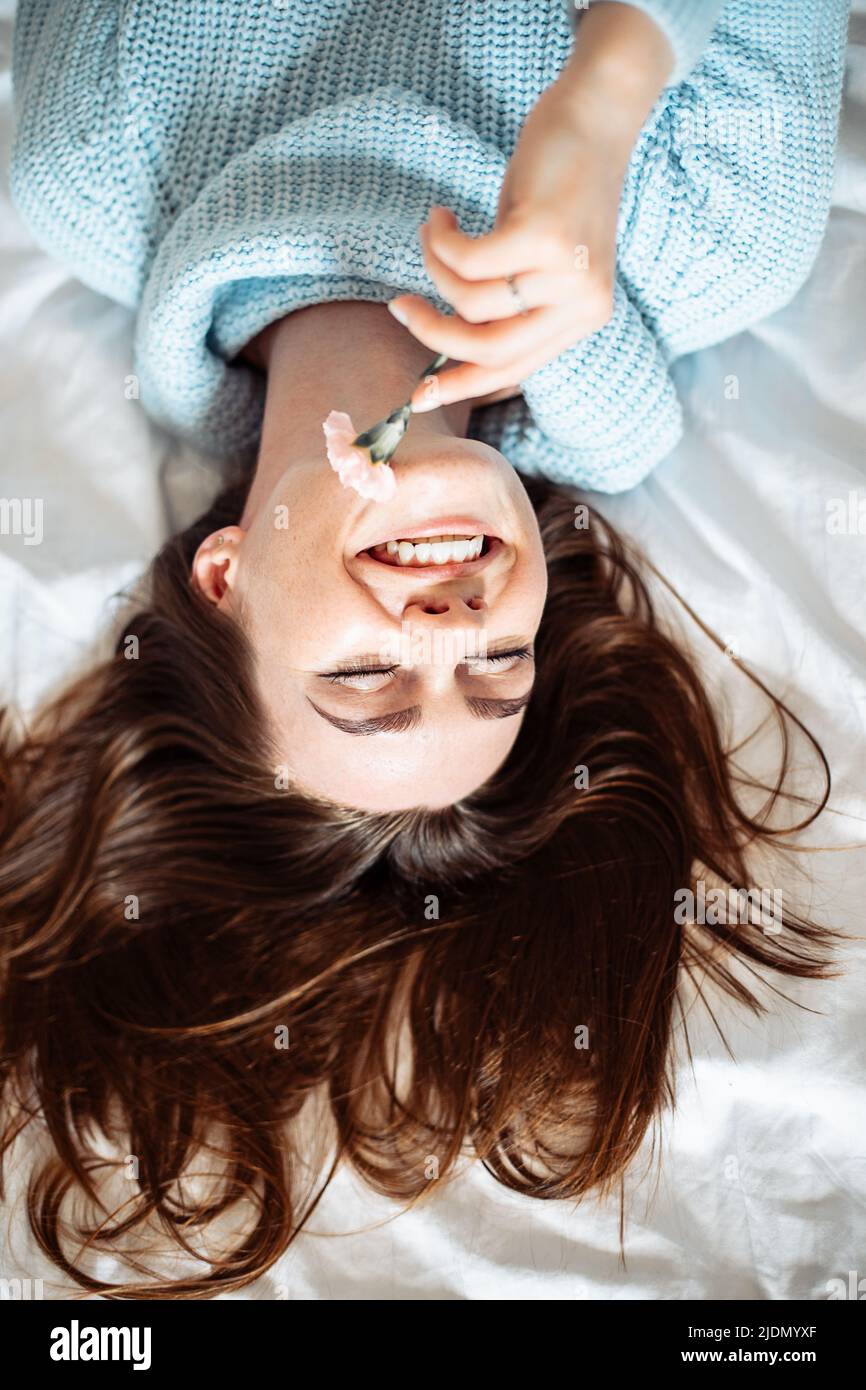 Ragazza carina in maglione blu con lunghi capelli scuri adagiati sul letto con fiori e divertenti Foto Stock