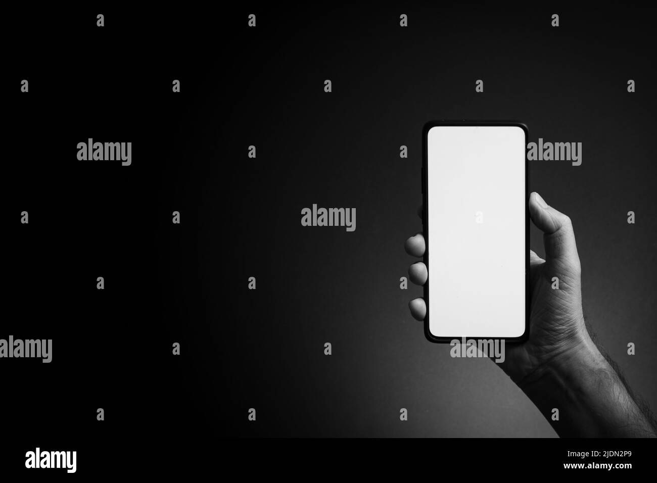 Immagine in bianco e nero della mano dell'uomo che tiene lo smartphone nero in posizione verticale con schermo bianco vuoto isolato su sfondo scuro con illuminazione straordinaria e. Foto Stock