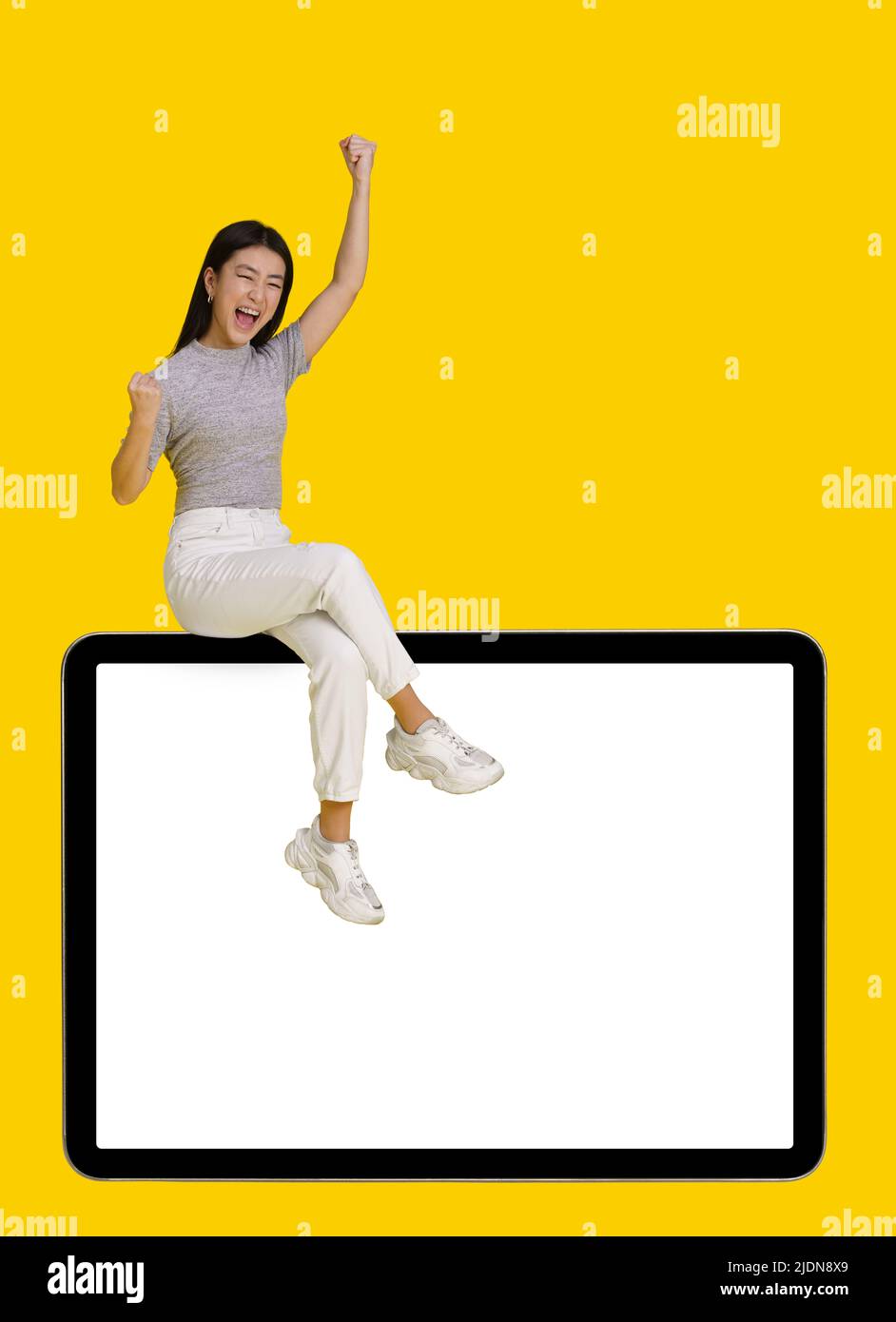 Vinci un gesto eccitato ragazza asiatica seduta su un enorme tablet pc digitale con schermo bianco isolato su sfondo giallo. Simulazione del posizionamento dei prodotti per la pubblicità tramite app mobile. Spazio di copia. Foto Stock
