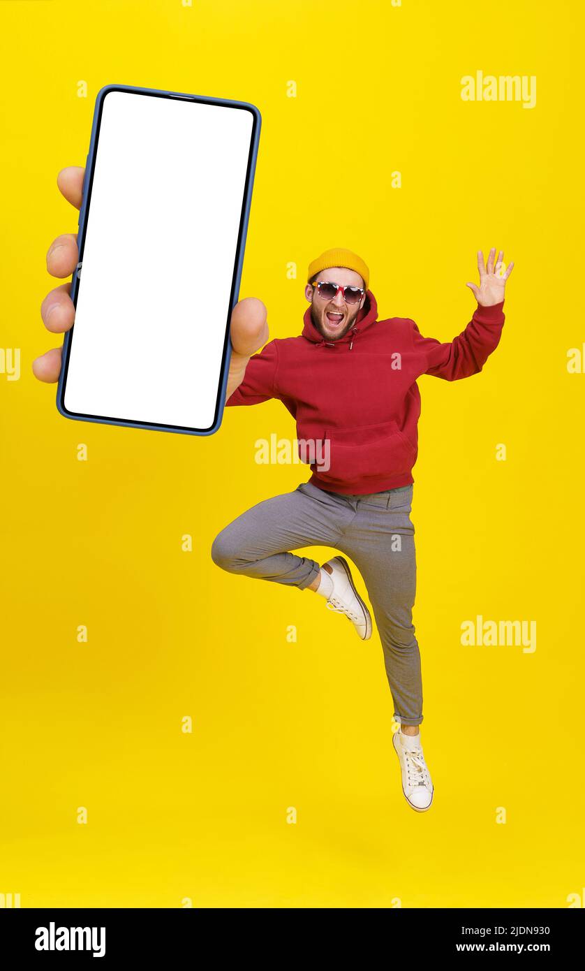 Giovane uomo in UK bandiera occhiali da sole saltare in gioia come balletto ballerino tenendo smartphone indossando casual felpa con cappuccio rosso e jeans isolato su sfondo giallo. Posizionamento dei prodotti pubblicitari tramite app mobile. Foto Stock