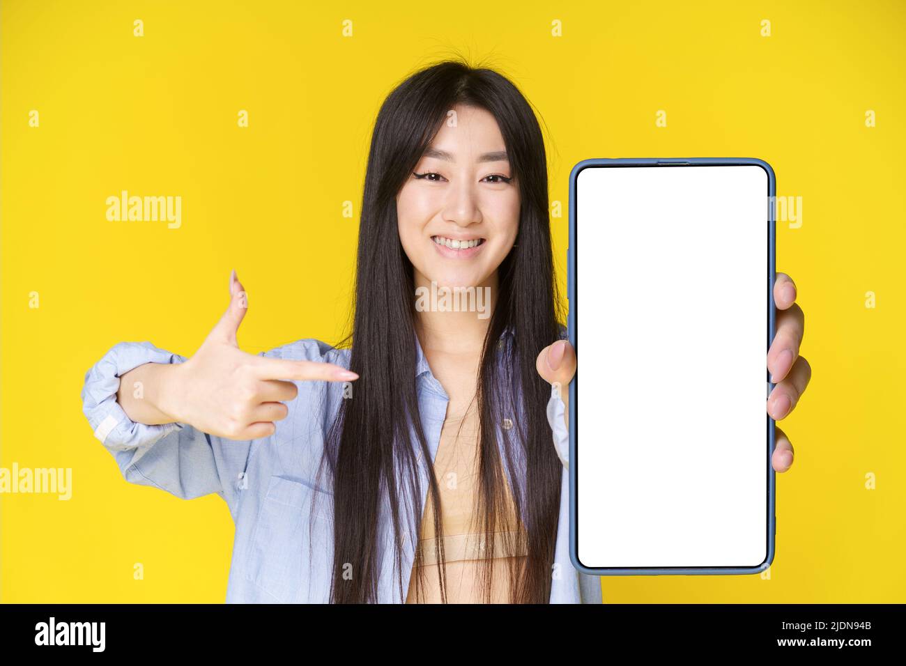 Ragazza asiatica affascinante con smartphone in mano puntando il dito su schermo bianco ed emozionante guardando la fotocamera isolato su sfondo giallo. Grande offerta. Posizionamento di prodotti pubblicitari per app mobili. Foto Stock
