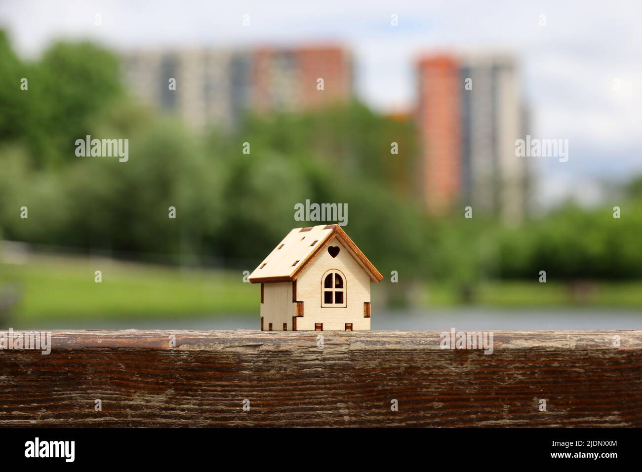 Modello di casa in legno sullo sfondo del lago estivo e alti edifici residenziali. Concetto di casolare di campagna, immobiliare in zona ecologica Foto Stock