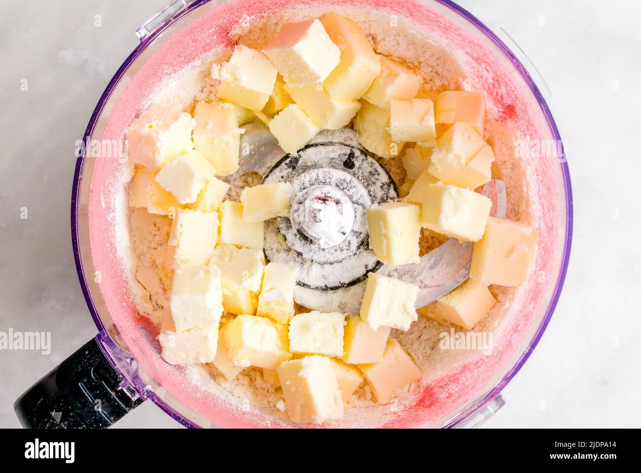 Preparazione della crosta di torta di fragole rosa in un robot da cucina: Vista dall'alto del burro a cubetti, delle fragole in polvere e della farina in un recipiente del robot da cucina Foto Stock