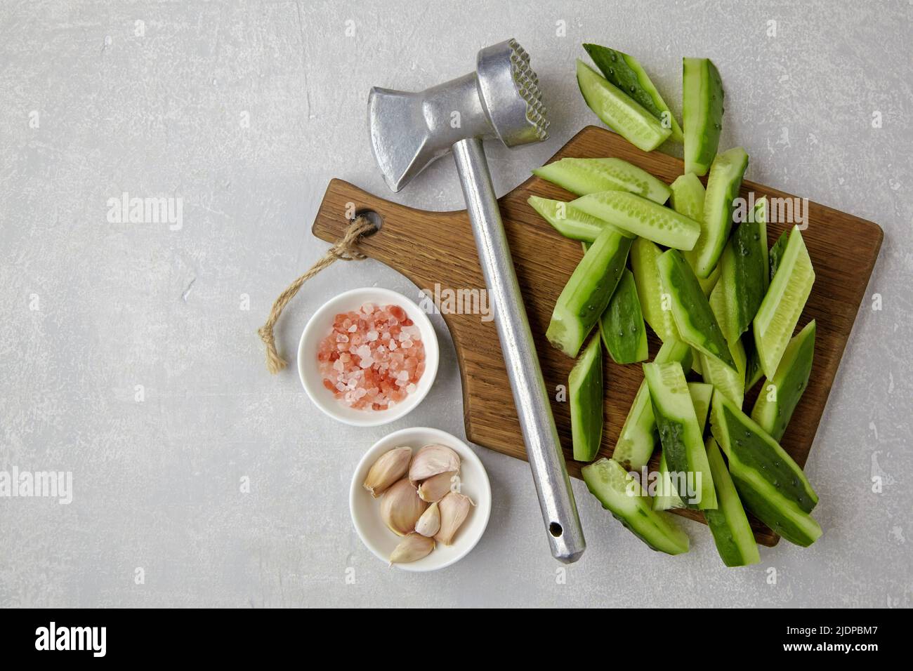 Tagliare cetrioli freschi su un tagliere di legno, martello da cucina, aglio e sale rosa su un tavolo di cemento leggero. Cucina di antipasti da cucum rotto Foto Stock