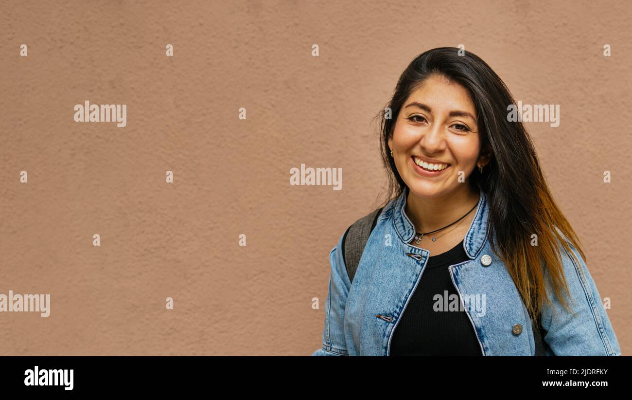 Un primo piano di una donna latina dai capelli lunghi che indossa la giacca jean e sorride guardando la fotocamera con sfondo unicolor dietro Foto Stock