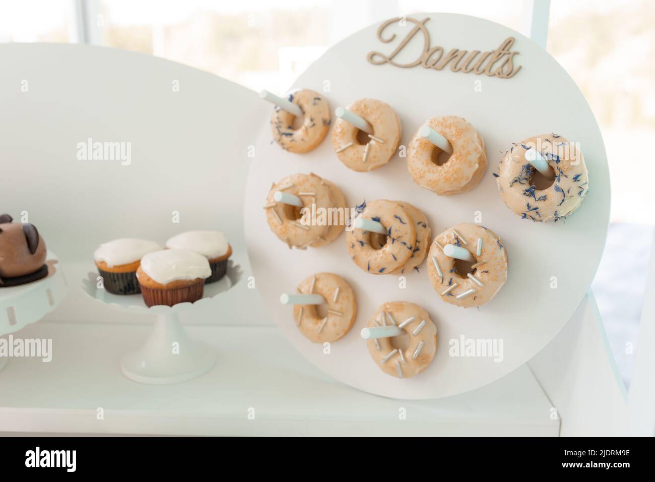 Serve ciambelle decorate con farcitura e spolverini di cioccolato e cupcake cremosi su vassoio, sfondo bianco. Dolce dessert, festa dell'evento Foto Stock