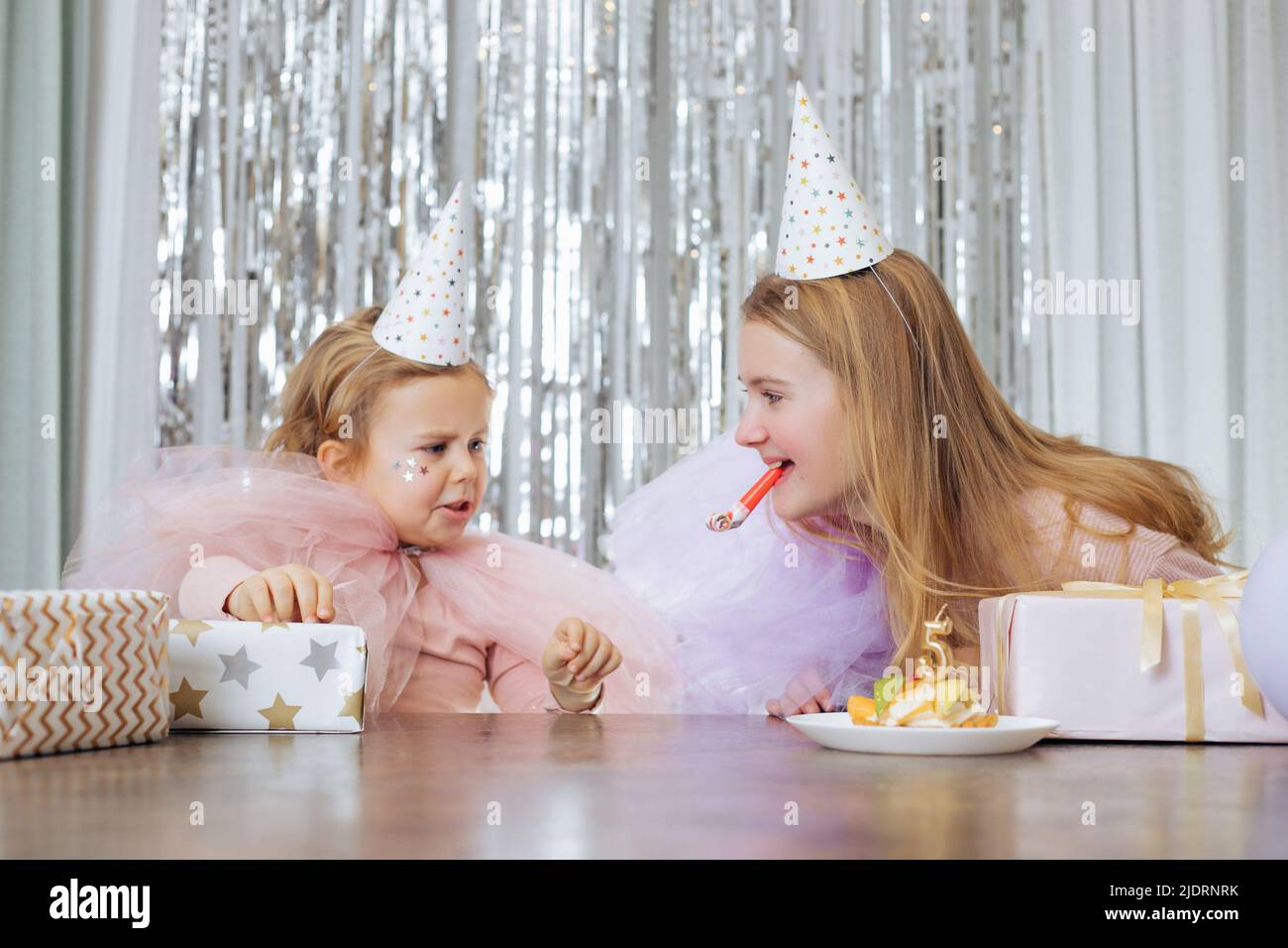 Emozioni dei bambini. ragazza di 5 anni in cappello da festa e abito festivo è sconvolta circa qualcosa alla sua celebrazione di compleanno, sua sorella più anziana sta provando Foto Stock