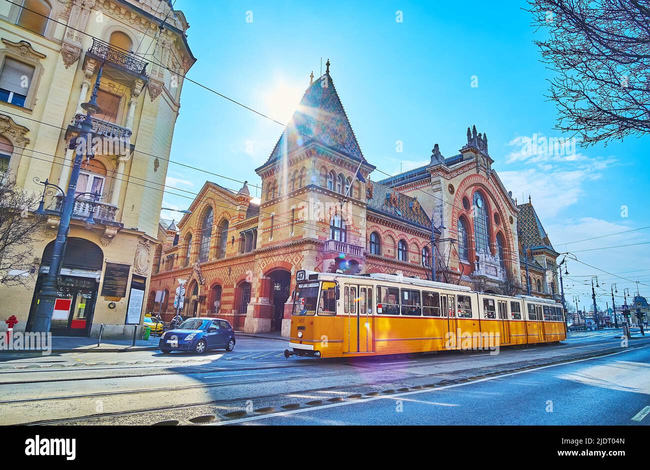 La scena urbana con giro in tram giallo d'epoca, la costruzione del mercato Centrale e il sole luminoso, splendente sul suo tetto, Budapest, Ungheria Foto Stock