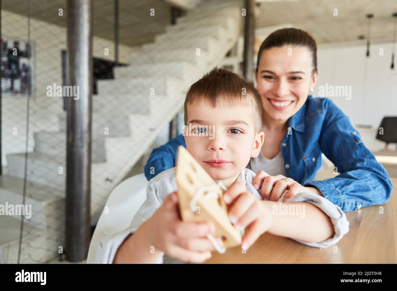 Il bambino mostra orgogliosamente un blocco di filettatura in legno come un motore abilità gioco educativo con la madre in background Foto Stock