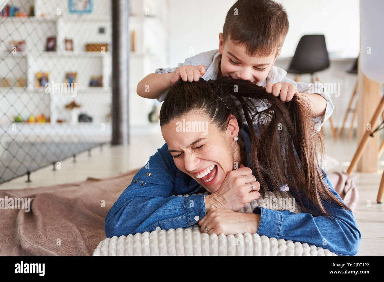 Ragazzo che tira i lunghi capelli della madre per divertirsi a casa nel soggiorno Foto Stock