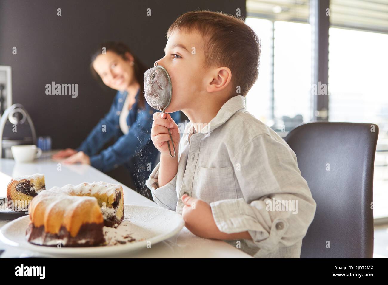 Il ragazzino lecca il setaccio con lo zucchero in polvere mentre cuoce la torta con la madre Foto Stock