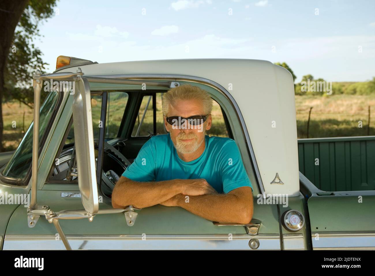 Uomo biondo (40-60 anni) con barba che si apre la finestra di un camion d'epoca pick up parcheggiato in una zona rurale. Foto Stock