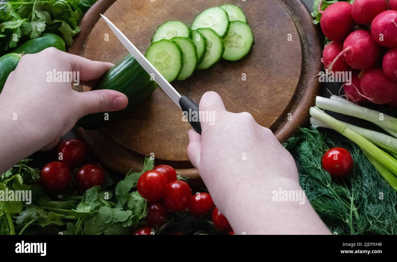 Tagliare a mano i cetrioli con un coltello in ceramica bianca su un tagliere rotondo in legno. Intorno al bordo ci sono verdure fresche mature: Pomodori, cipolle, rad Foto Stock