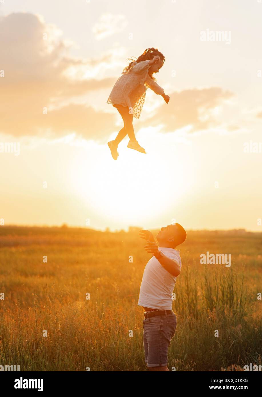 Felice padre con bambino ha un divertimento, gioca e getta sua figlia su prato contro cielo sfondo al tramonto. Foto Stock
