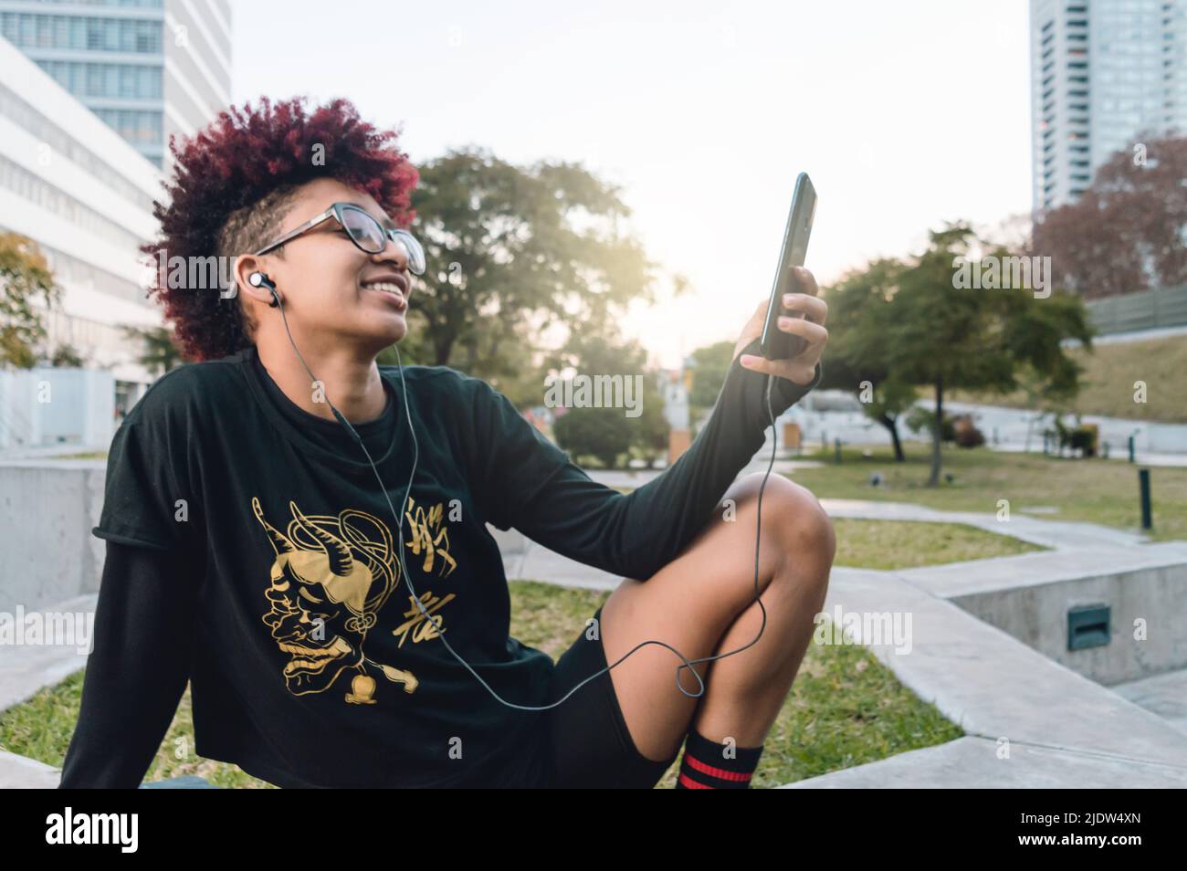 giovane bruna ispanica donna latina, colombiana con afro e occhiali, seduto all'aperto sorridendo rilassato nel parco intrattenuto guardando la rete sociale Foto Stock