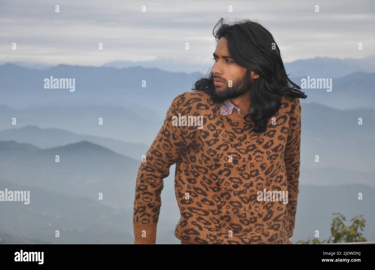 Un giovane indiano dai capelli lunghi e bearded che guarda lateralmente mentre si alza sullo sfondo delle montagne Foto Stock