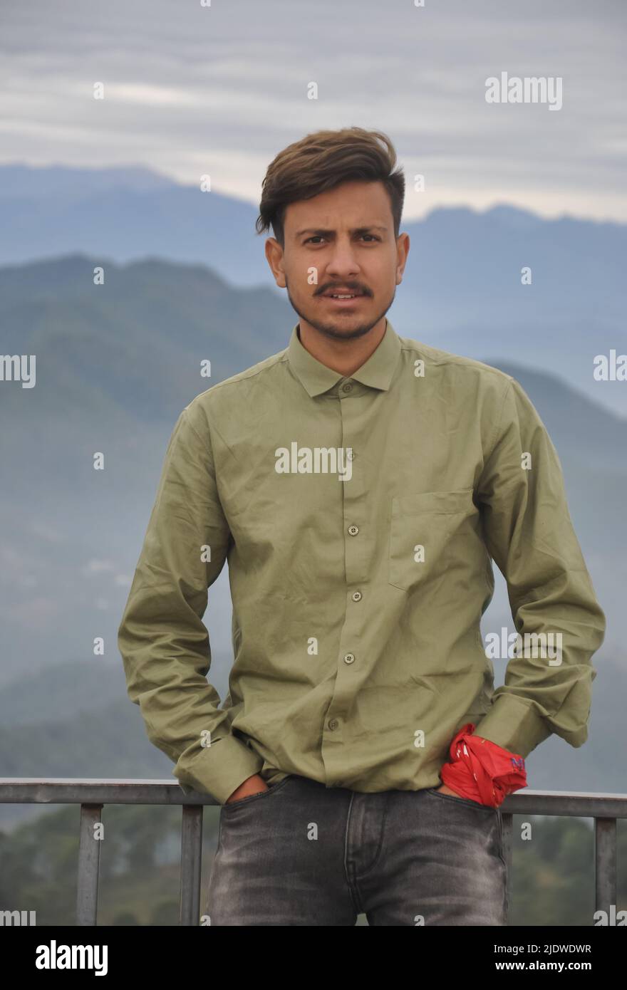 Un giovane indiano affascinante che guarda la macchina fotografica con le mani in tasca mentre si trova sullo sfondo delle montagne Foto Stock