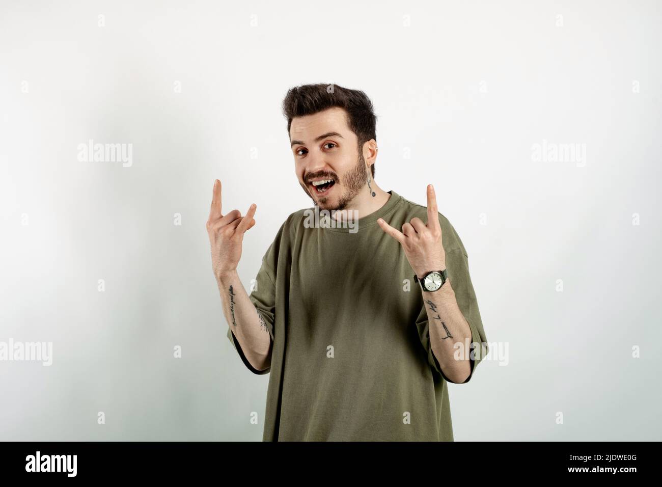 Felice uomo giovane che indossa la t-shirt verde posa isolato su sfondo bianco facendo rock simbolo con le mani in su con espressione pazza. Star della musica. Pesante c Foto Stock