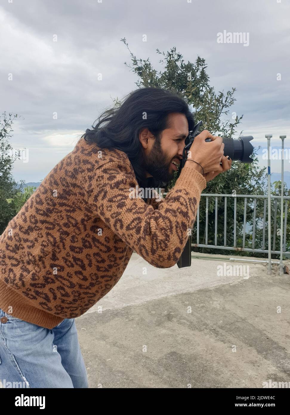 Vista laterale di un giovane indiano con capelli lunghi e bearded che scatta foto con la macchina fotografica mentre si alza all'esterno Foto Stock