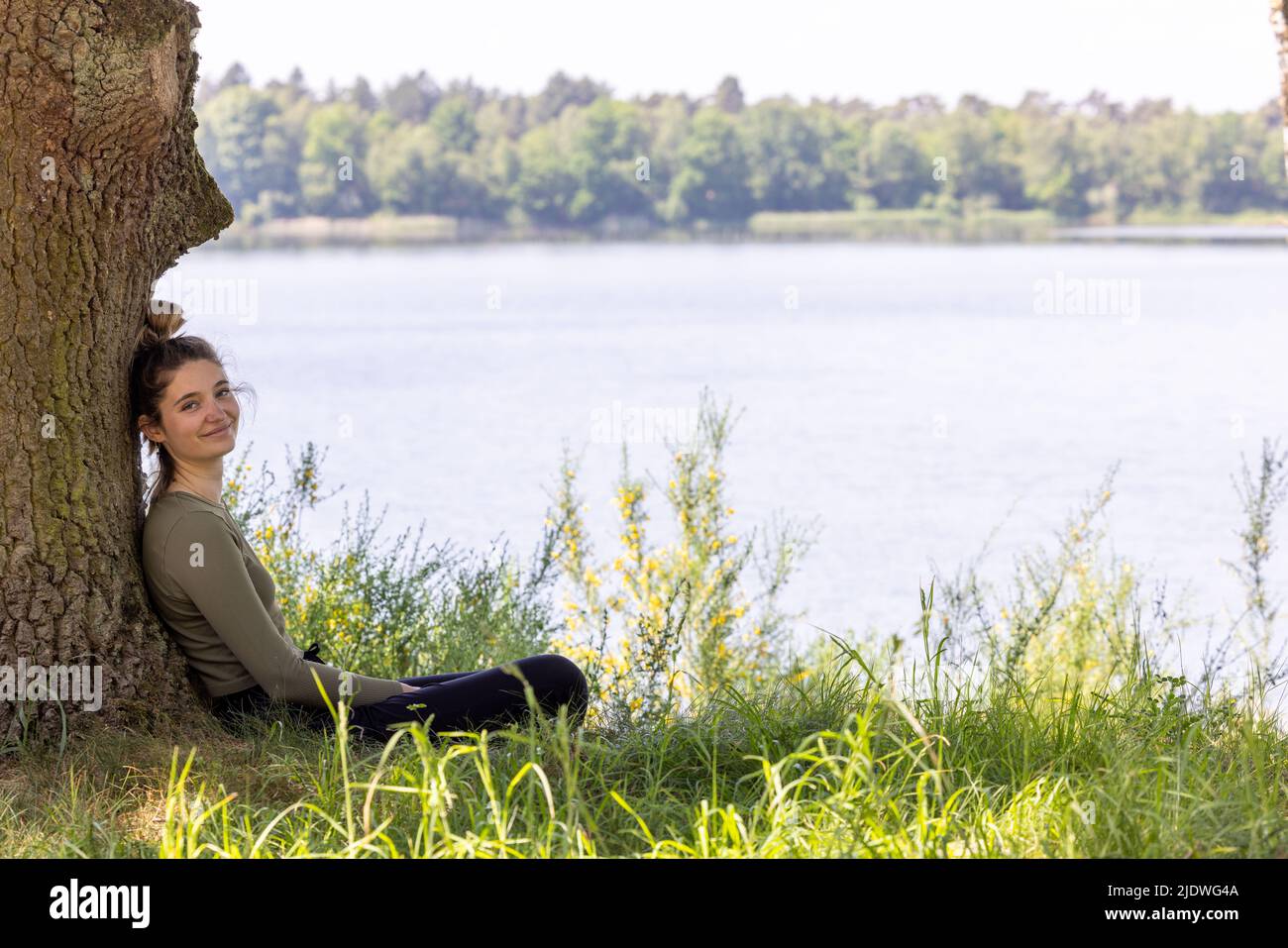 Giovane donna bruna seduta in foresta verde gode il silenzio e la bellezza della natura guardando su un lago di foresta blu in una giornata estiva. Foto di alta qualità Foto Stock