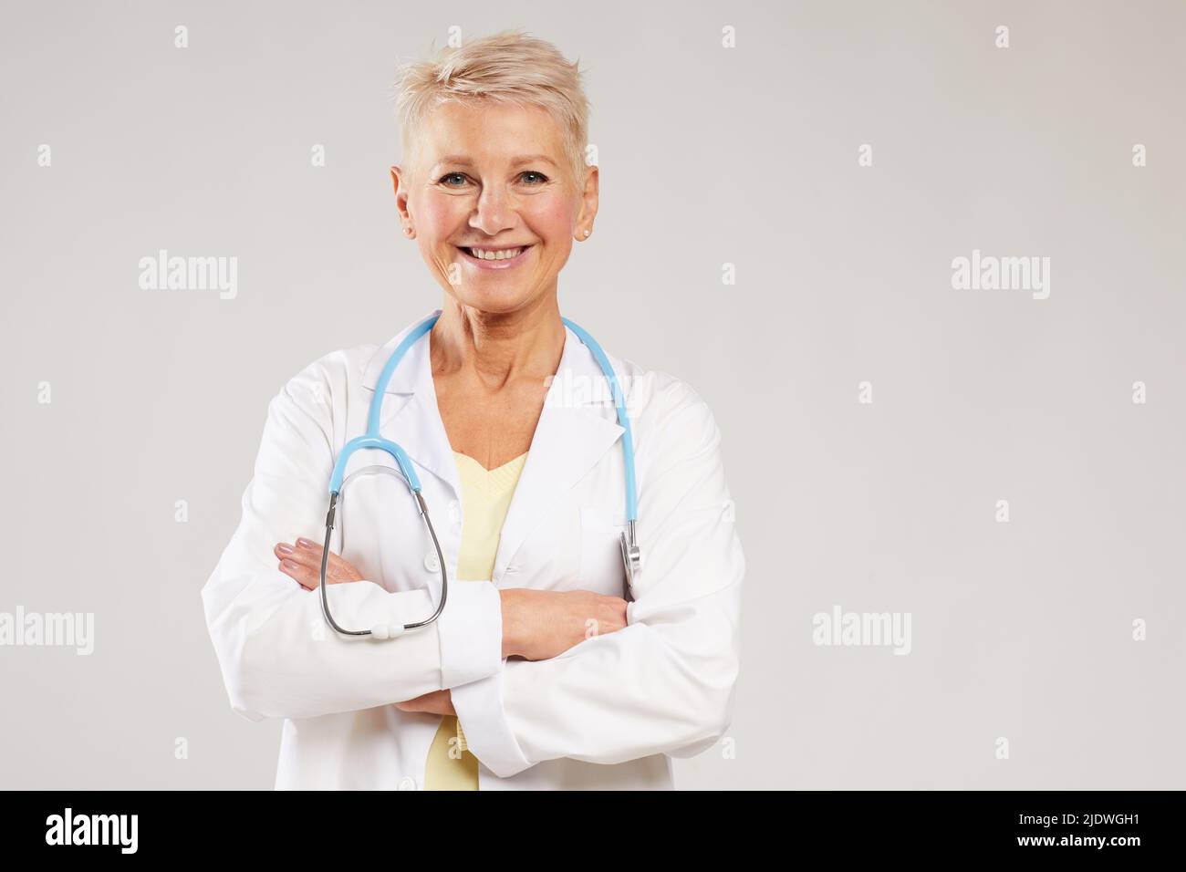 Ritratto di medico generico femminile maturo positivo con stetoscopio in piedi su sfondo bianco e incrociando le braccia sul petto Foto Stock