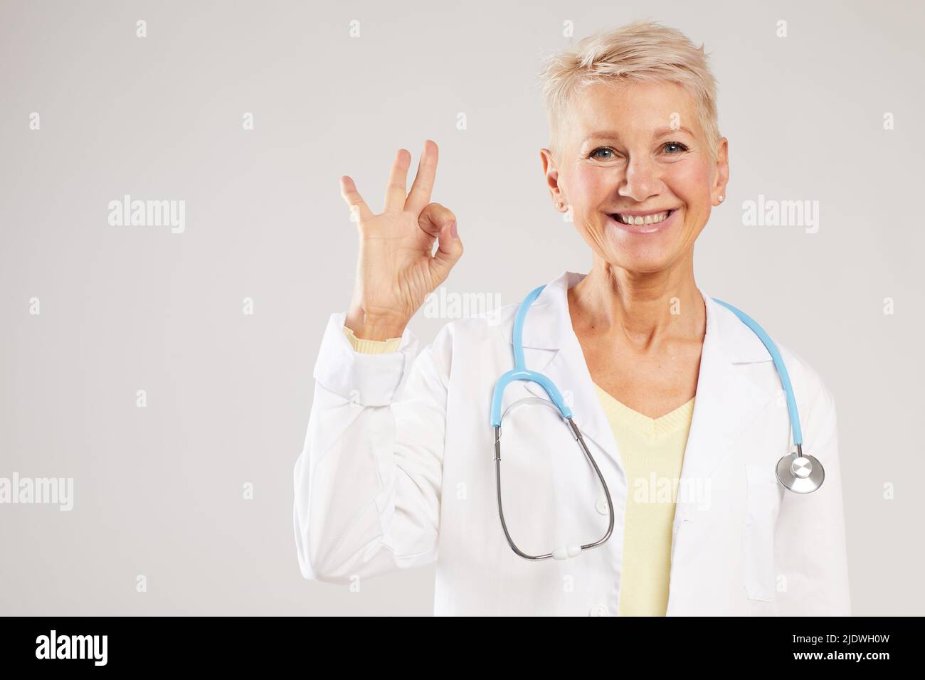 Ritratto di medico specialista femminile positivo centro con stetoscopio sul collo in piedi su sfondo bianco e mostrando segno ok Foto Stock