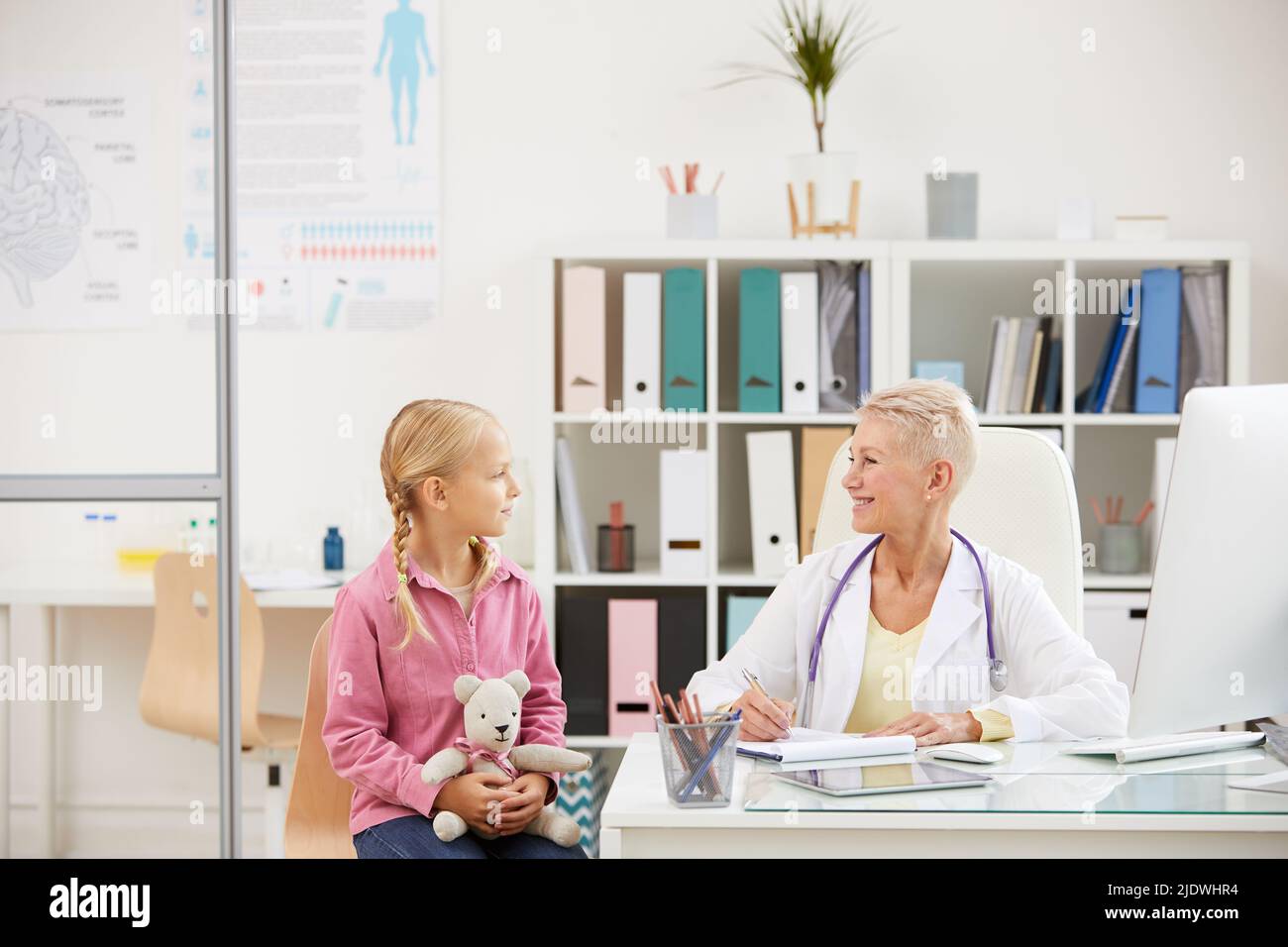 Positivo attraente pediatra femminile con stetoscopio seduto al tavolo e riempire la storia medica della ragazza durante l'appuntamento in ufficio Foto Stock