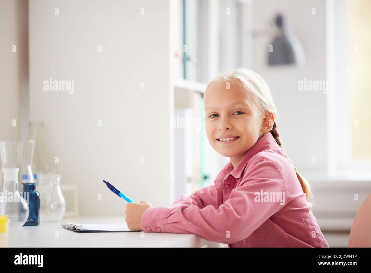 Ritratto di sorridente bella scolaretta con bionda trecce seduti a tavola in laboratorio e facendo note sull'esperimento Foto Stock