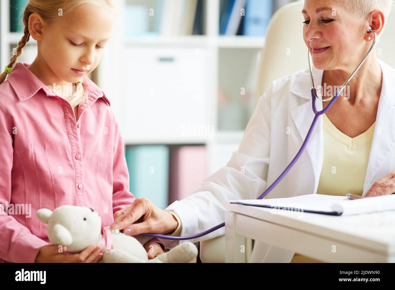 Medico positivo bambini in camice da laboratorio utilizzando stetoscopio mentre ascolta l'orso giocattolo, lei giocando con piccolo paziente durante la consultazione Foto Stock