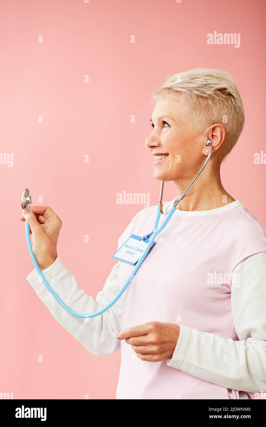 Positivo bel medico maturo biondo-capelli in uniforme rosa e bianco in piedi su sfondo rosa tenue e con stetoscopio Foto Stock