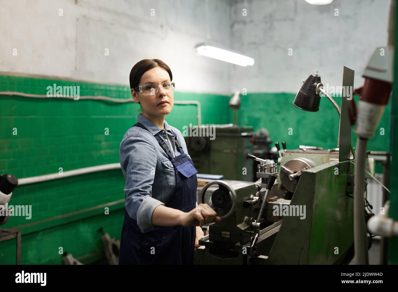 Ritratto di fidato ingegnere femminile attraente in occhiali di sicurezza utilizzando il tornio manuale mentre si lavora con pezzi di metallo Foto Stock