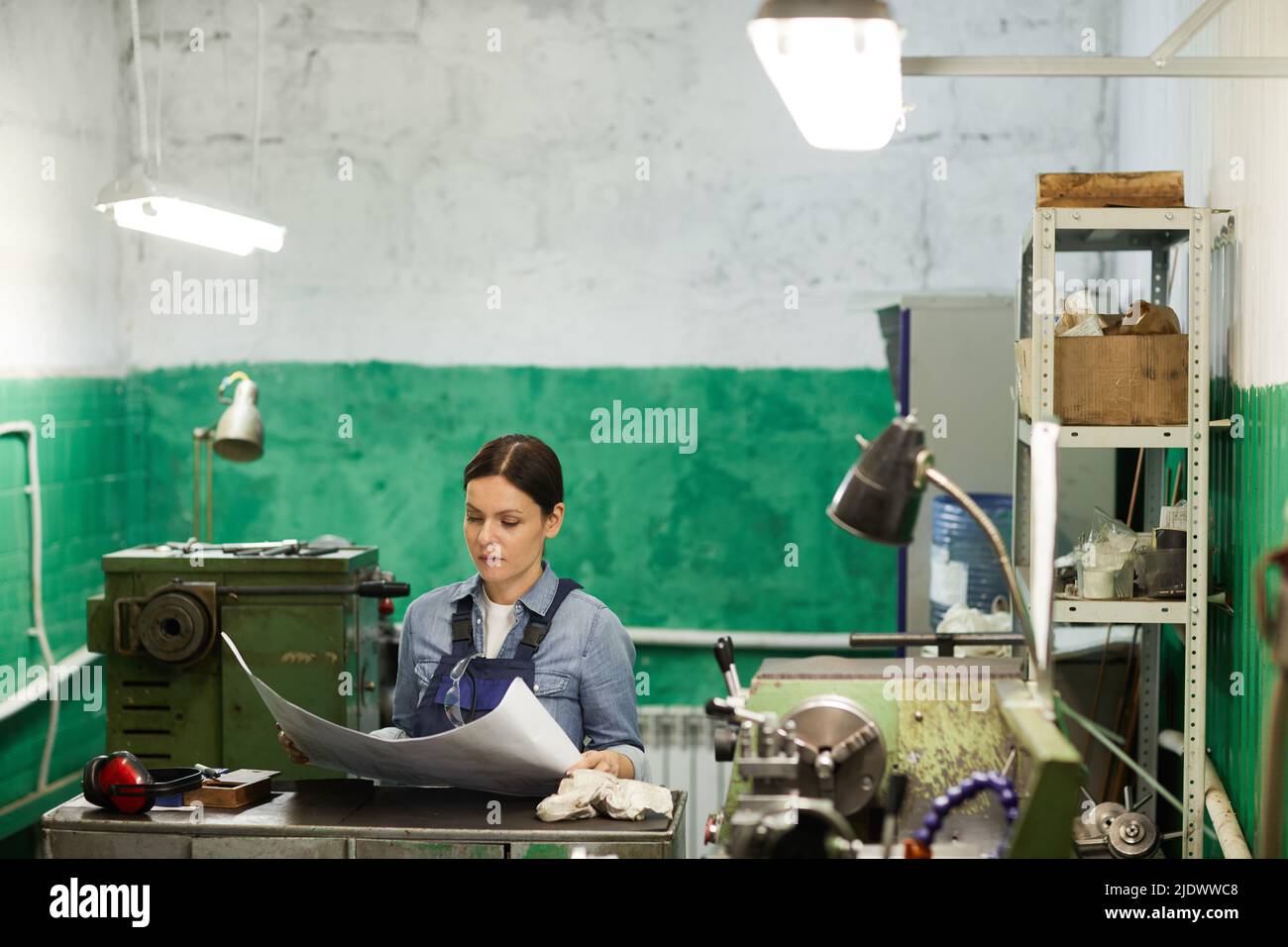 Pensierosa ingegnere di mezza età in abbigliamento da lavoro in piedi al banco di metallo e leggere il disegno tecnico in negozio industriale Foto Stock
