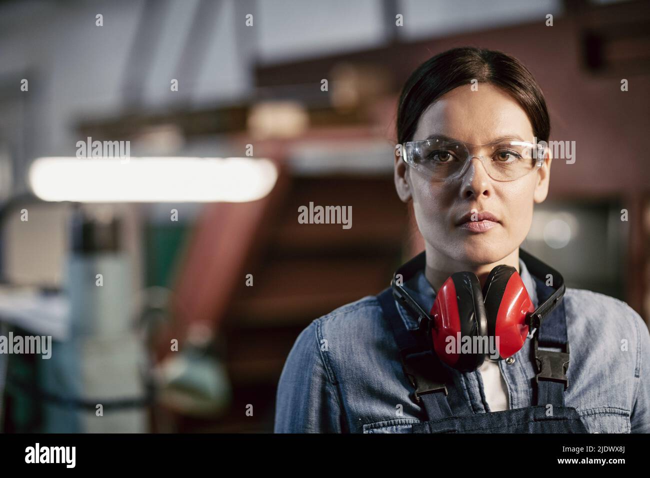 Ritratto di bruna bruna grave e ruvida di mezza età donna in occhiali di sicurezza e protezioni per le orecchie che lavorano presso impianti industriali Foto Stock