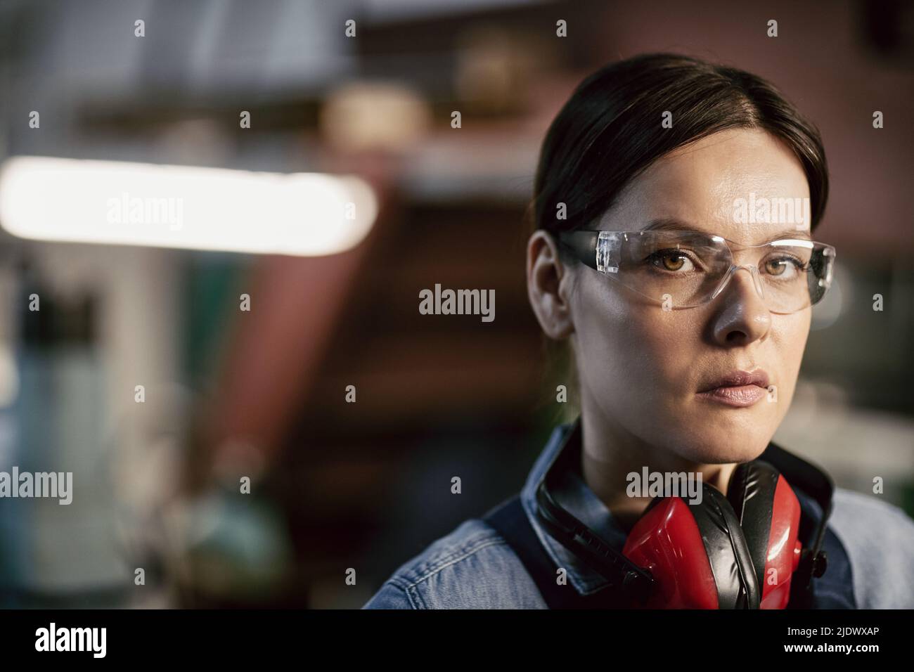 Ritratto di donna seria e attraente resistente con capelli marroni che indossano protezioni per le orecchie rosse sul collo in piedi presso l'officina di fabbrica Foto Stock
