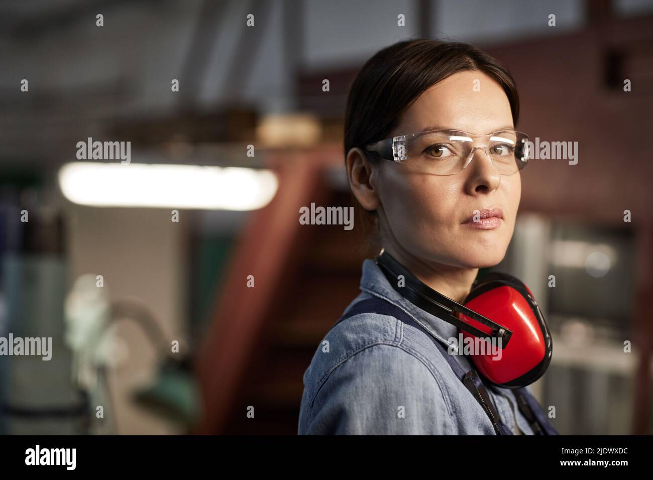Ritratto di fidata bella donna in occhiali di sicurezza e protezioni per le orecchie sul collo lavorando in fabbrica industriale Foto Stock