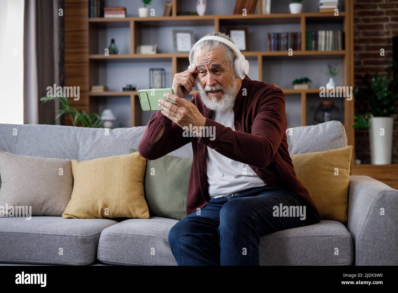 Felice uomo anziano dai capelli grigi in cuffie bianche che tengono lo smartphone e ridono. Vecchio uomo seduto sul divano a casa guardando contenuti divertenti Foto Stock