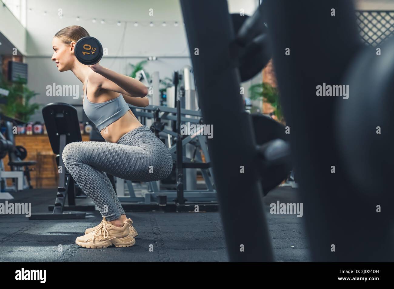 Scatto interno orizzontale di una ragazza caucasica bella vestita che si esercita in una palestra ben attrezzata, sollevando un barbell di 10 chilogrammi. Foto di alta qualità Foto Stock