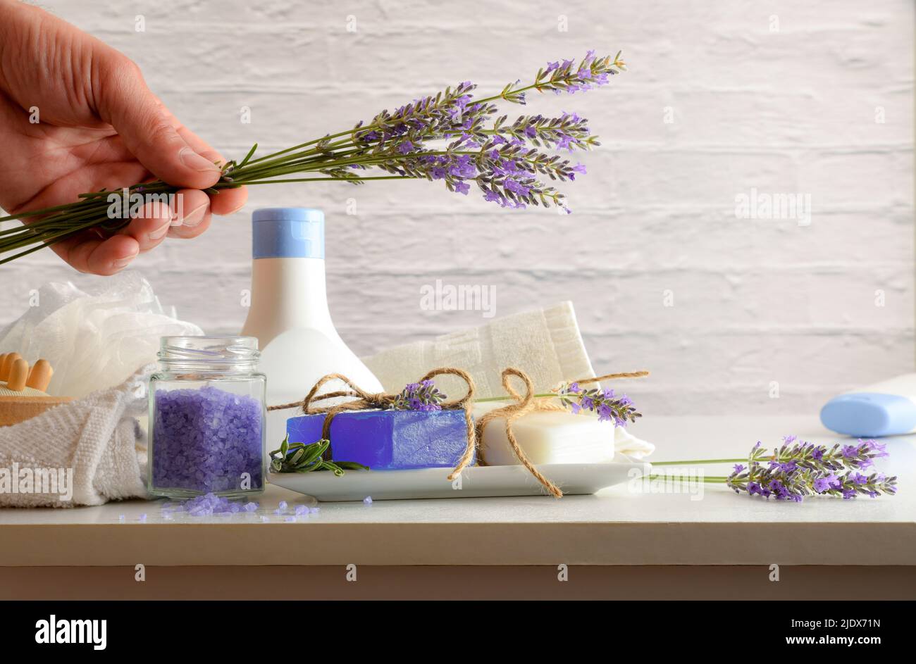 Prodotti per doccia con estratto di lavanda con elementi per l'igiene personale su tavola bianca e mano con bouquet. Vista frontale. Composizione orizzontale. Foto Stock