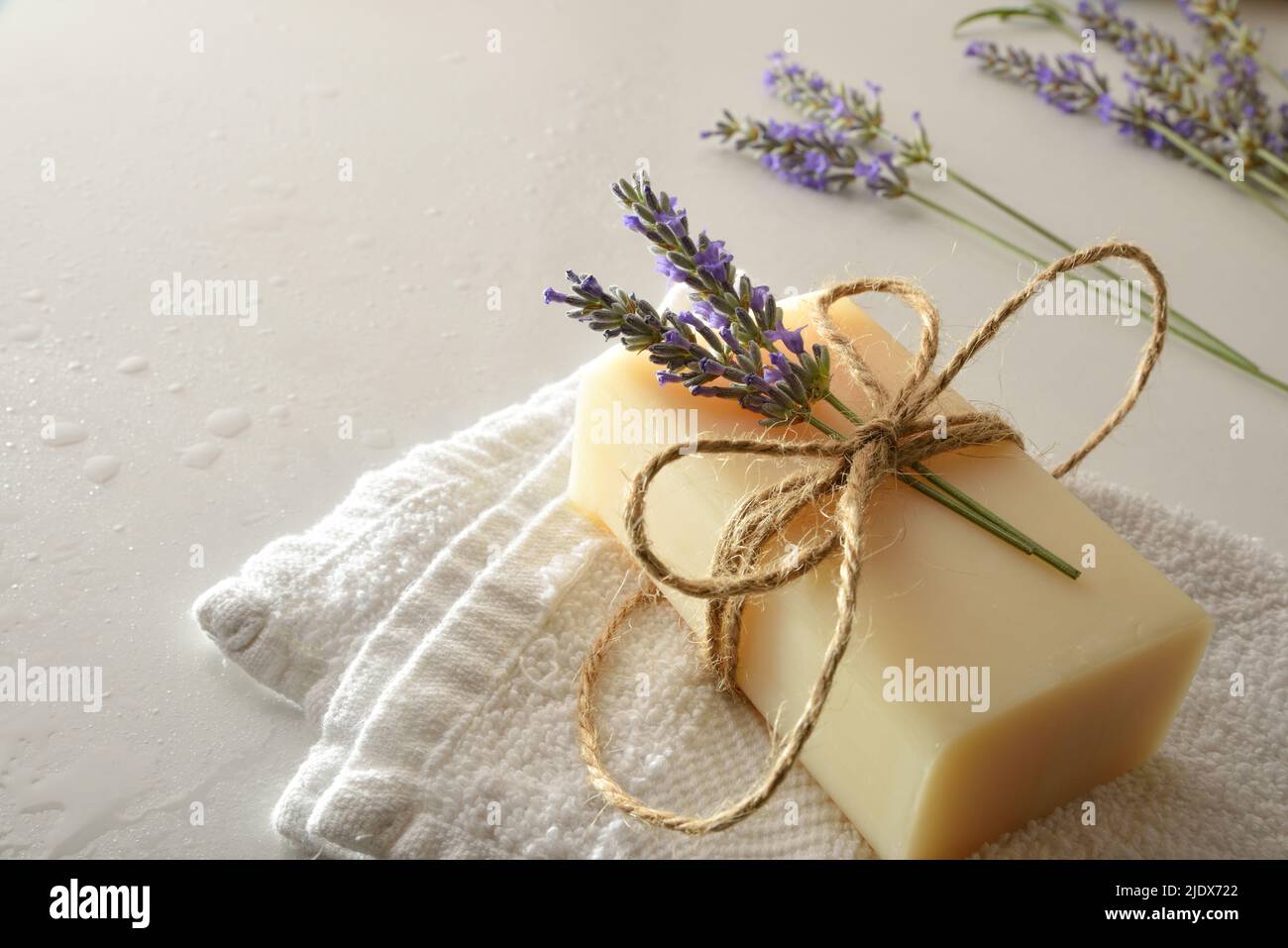 Dettaglio di sapone naturale con estratto di lavanda su tovagliolo su tavola bianca con gocce d'acqua e punte di lavanda in fiore. Vista rialzata. Horizonta Foto Stock