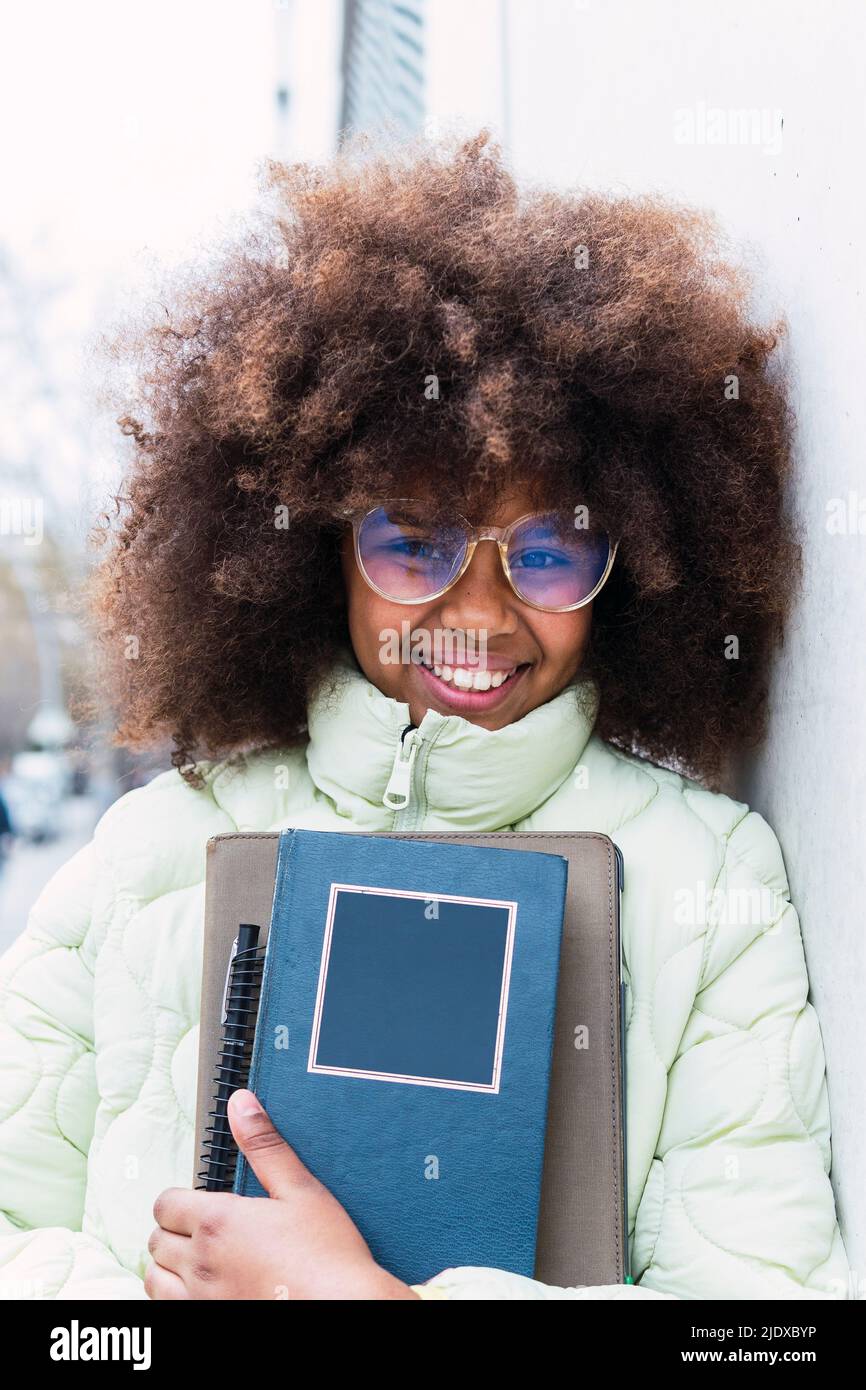Sorridendo Afro scolgirl con occhiali che tengono libri appoggiati sul muro Foto Stock