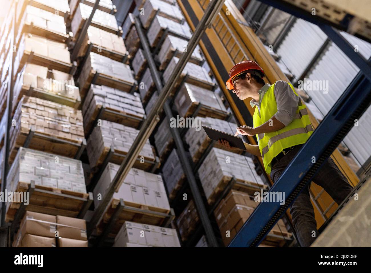 Lavoratore che indossa un elmetto che controlla l'inventario in piedi con un tablet PC tramite ringhiera in magazzino Foto Stock