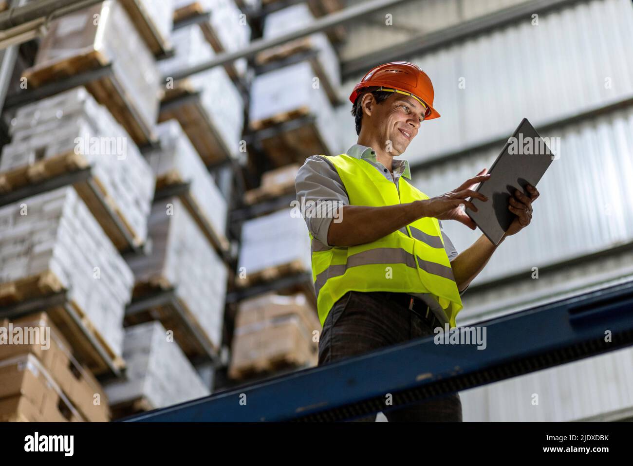 Sorridente lavoratore che indossa l'hardhat utilizzando un tablet PC da ringhiera in magazzino Foto Stock