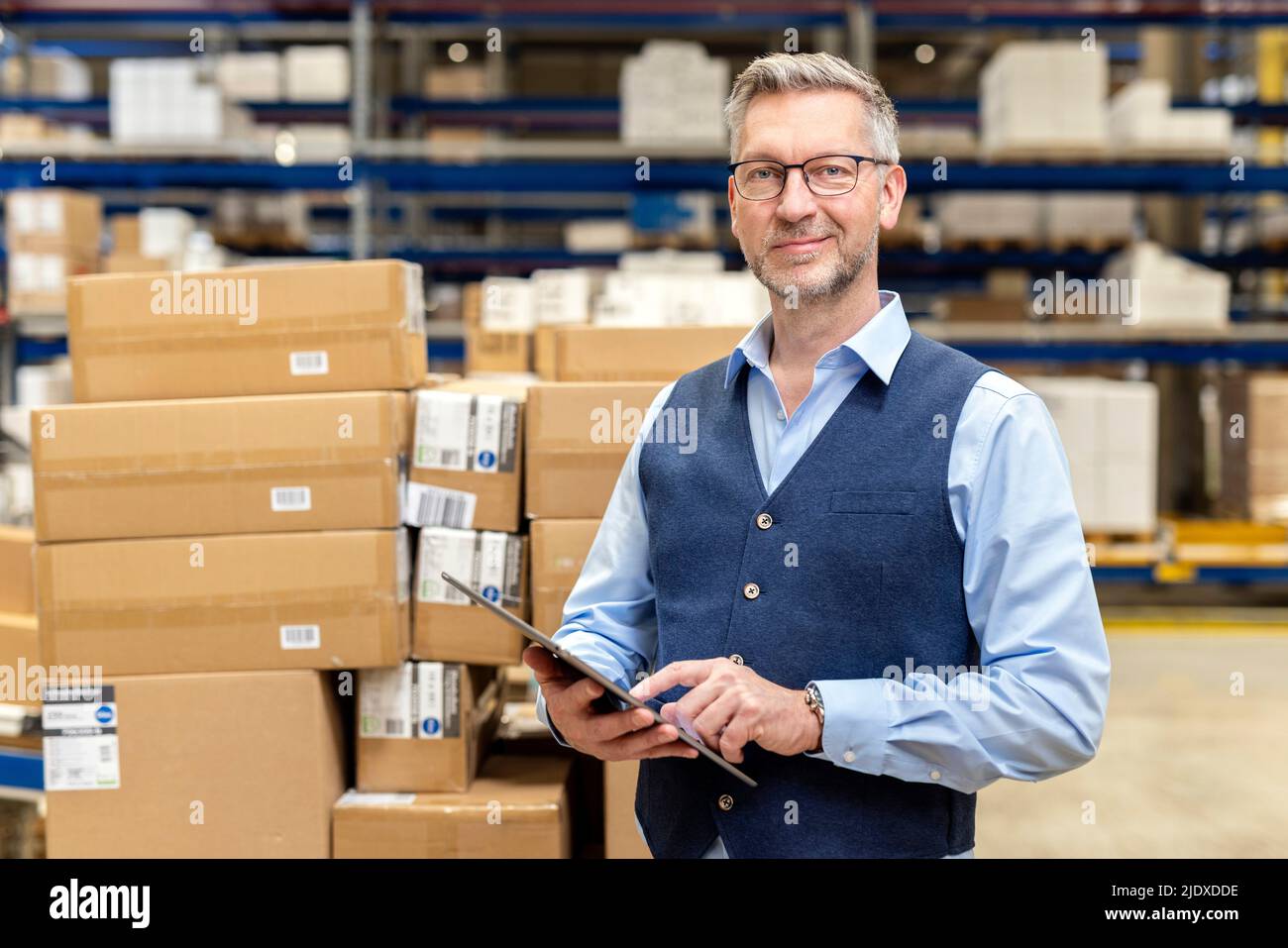 Un manager sorridente che indossa occhiali in piedi con un tablet PC davanti alle scatole di cartone in magazzino Foto Stock