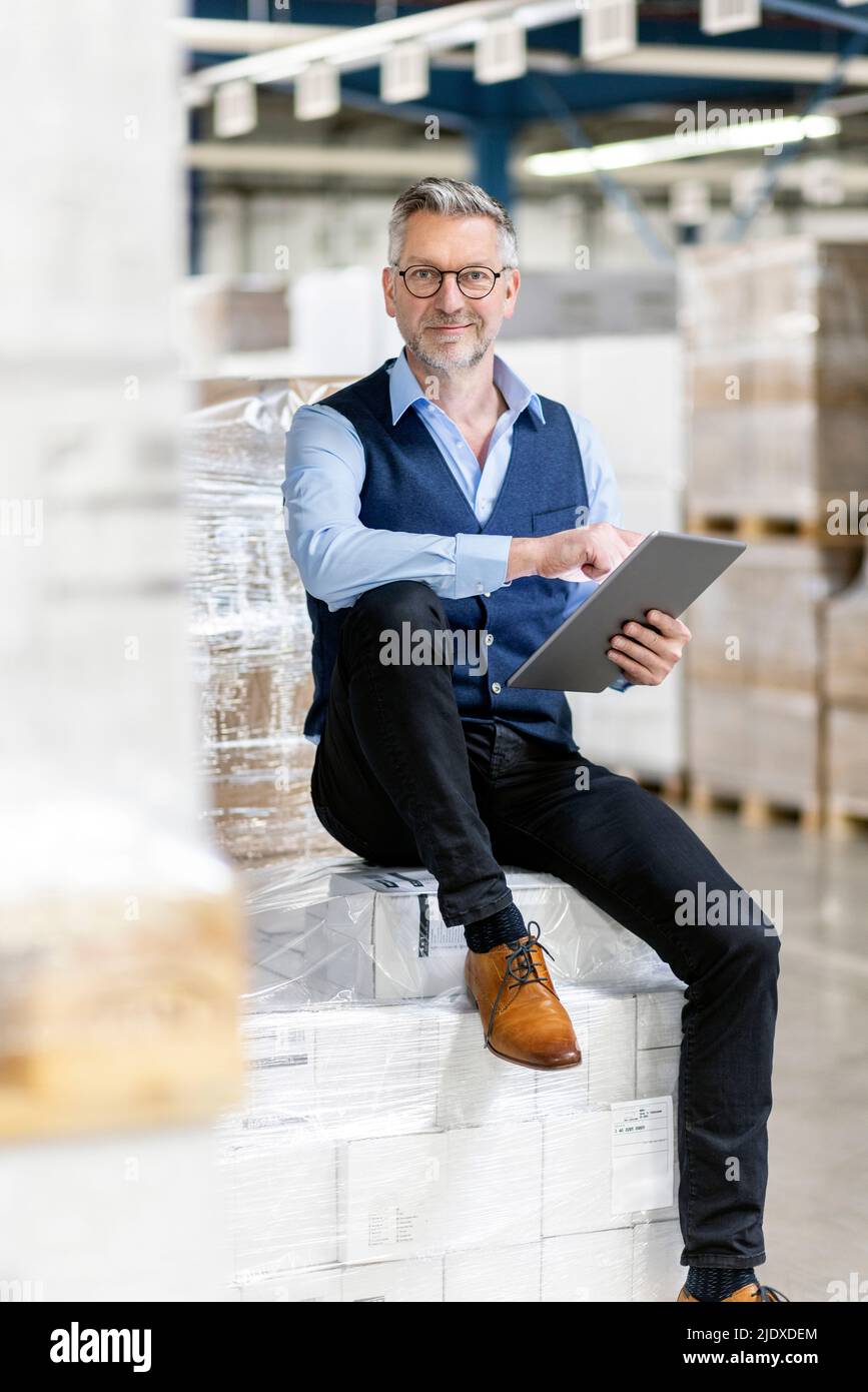 Un manager sorridente con un tablet PC seduto su scatole in magazzino Foto Stock